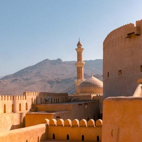 The walls and round tower of Nizwa Fort, with a mosque and mountains behind