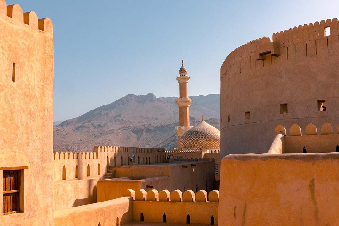 The walls and round tower of Nizwa Fort, with a mosque and mountains behind