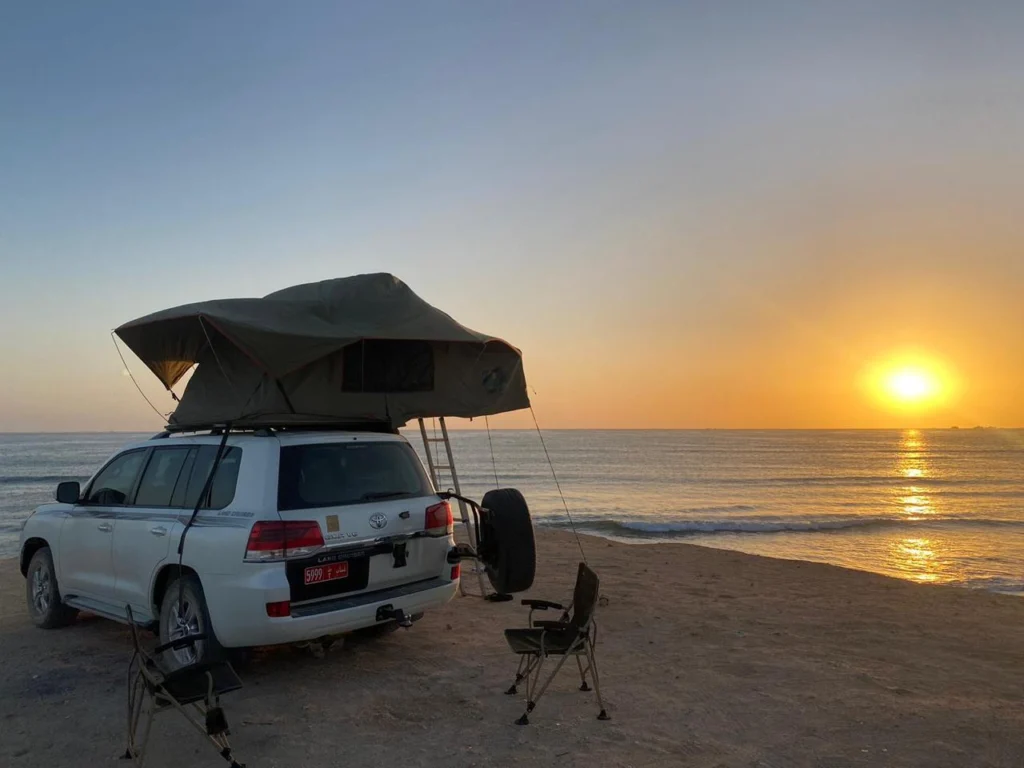 A white 4x4 with its rooftop tent open on the beach at sunset, with camp chairs and the sun on the sea