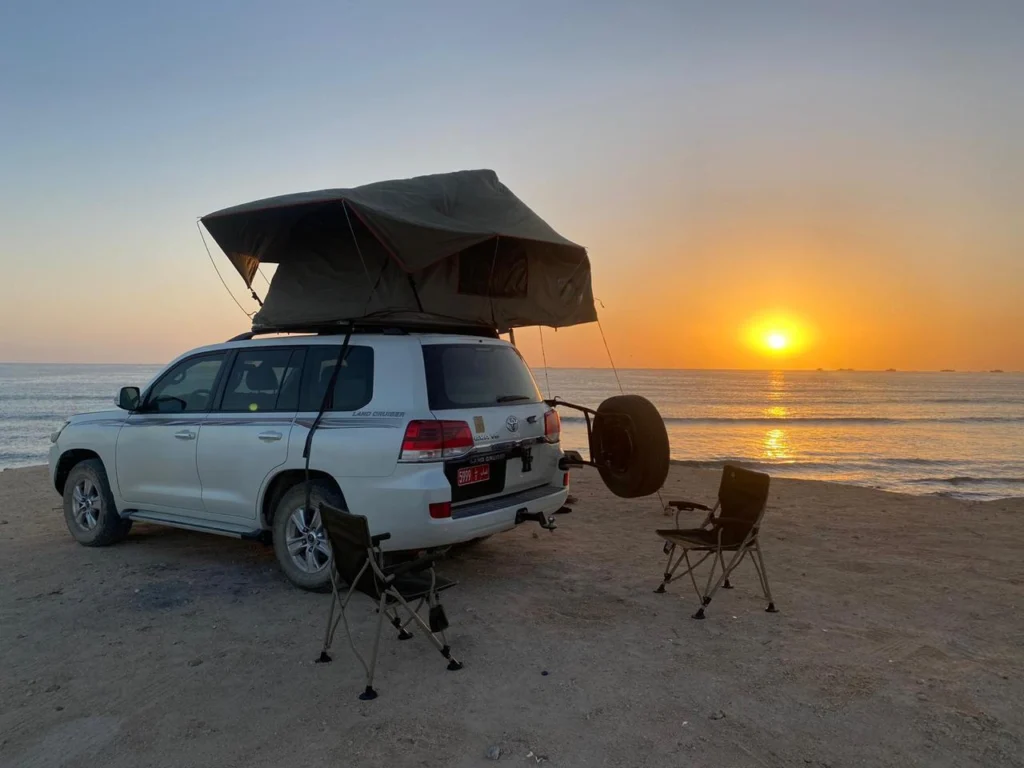 A white 4x4 with its rooftop tent open beside the sea at sunset, with a spare wheel on the back
