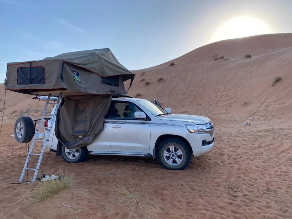 A rooftop-tent 4x4 on a dune with the low sun behind it and a ladder down from the tent