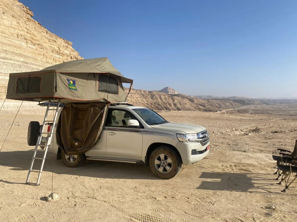 A 4x4 with its rooftop tent open on open ground below a pale desert cliff