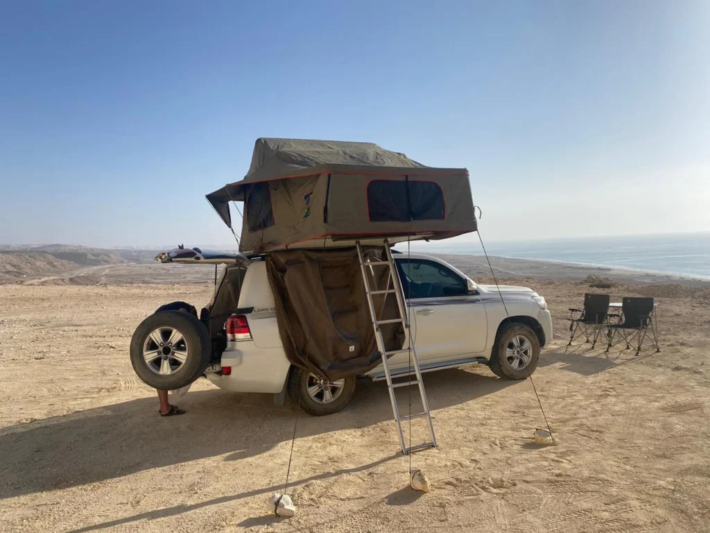 A 4x4 with its rooftop tent open on a gravel shore beside the sea, with camp chairs