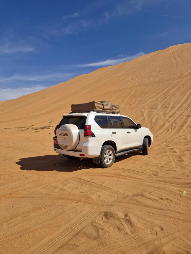 A white 4x4 with a roof rack on a steep sand dune under a blue sky