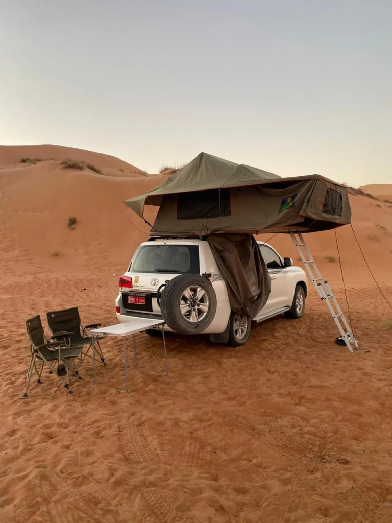 A white 4x4 with its rooftop tent open and camp chairs on desert sand