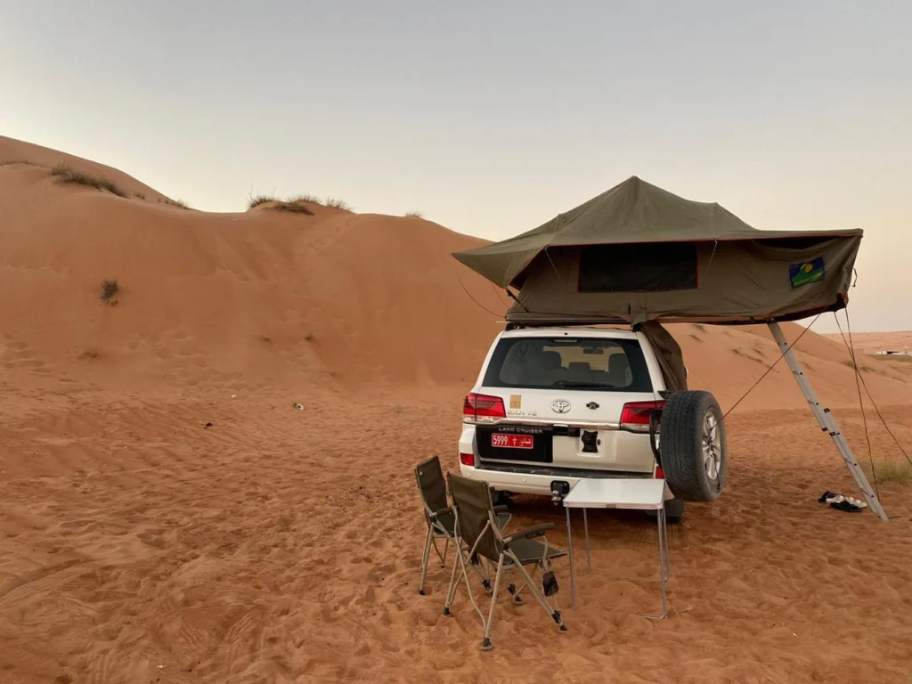 A white 4x4 with its rooftop tent open and camp chairs beside it, on desert sand at dawn