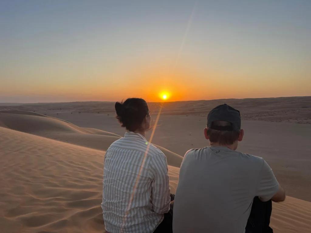 Two guests sitting on a dune watching the sun set over the desert